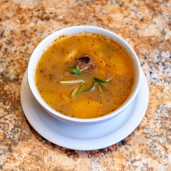 A bowl of Jamaican soup with meat, vegetables, and herbs on a speckled countertop.